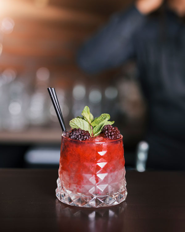 Red cocktail with blackberries and mint on top of a bar top